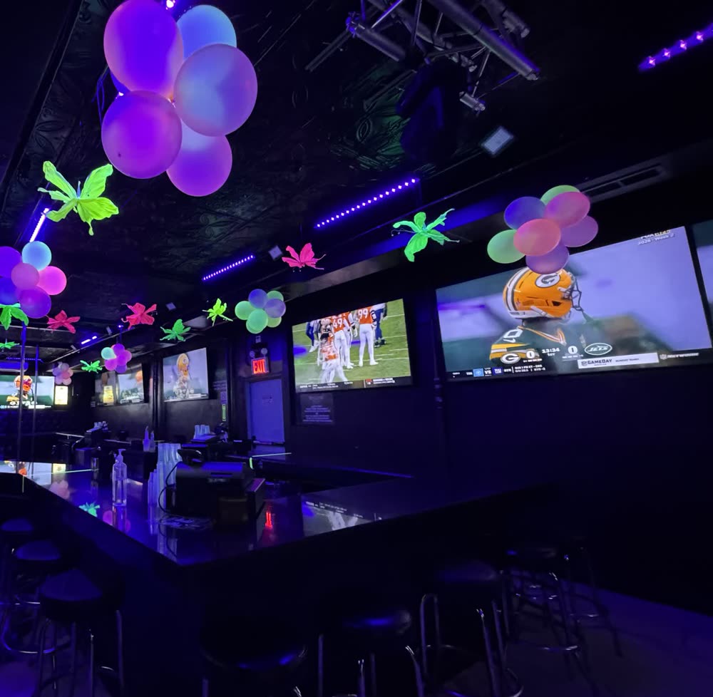 La sala de Sweets of NY: una barra larga con bancos, pantallas grandes encendidas encima, el escenario con el tubo a un lado y luces de colores en la estructura del techo.